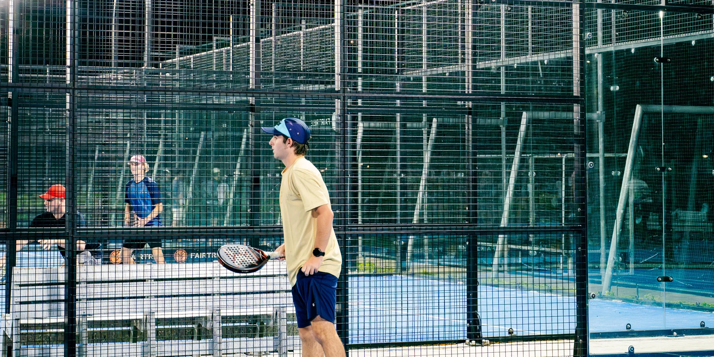 A player waiting at the fence under the floodlights, racket in hand, spectators watching from the next court