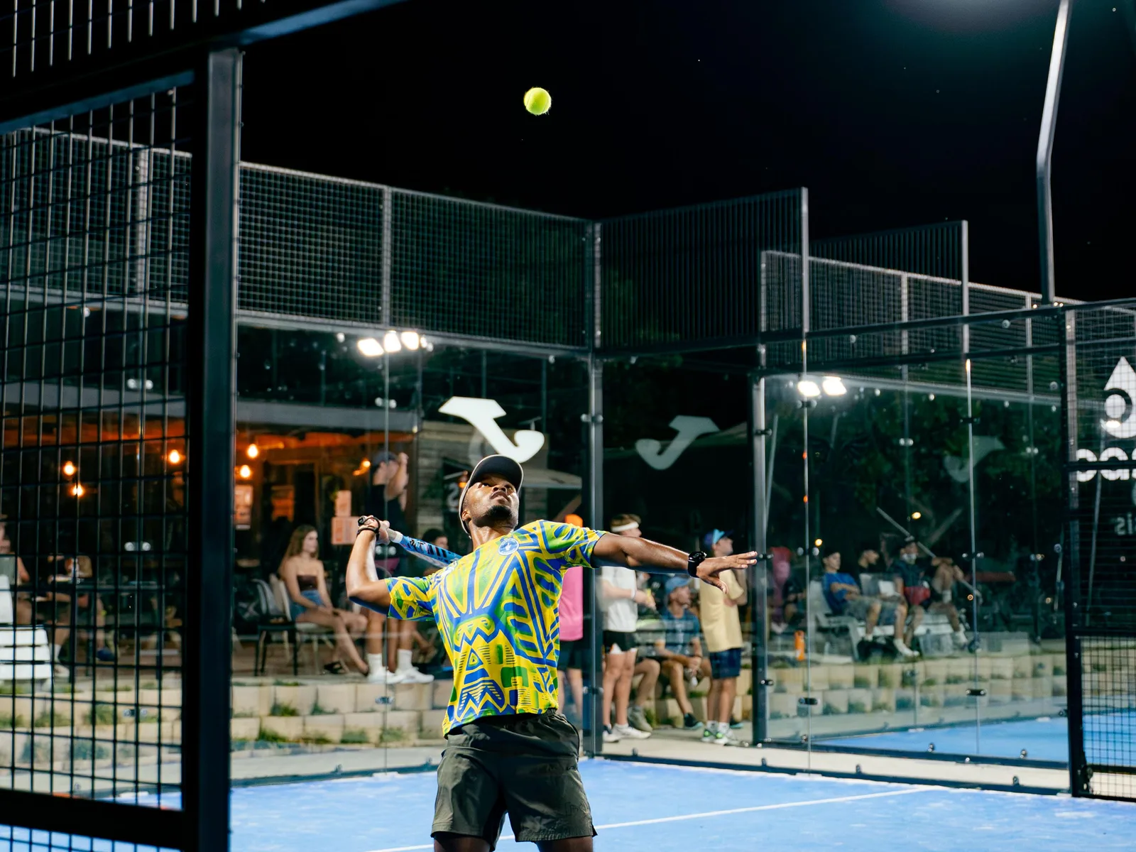 A player stretching into a forehand under the floodlights, the ball in the air beside him